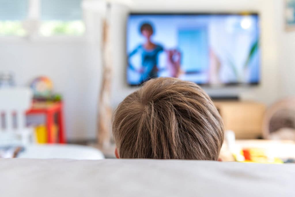 Die Kinderpraxis Ein Kind mit hellbraunem Haar sitzt in einem hellen, modernen Wohnzimmer auf einer Couch und blickt auf einen Fernseher, auf dem das verschwommene Bild einer Frau zu sehen ist. Kinderarzt Braunschweig Schlosscarree