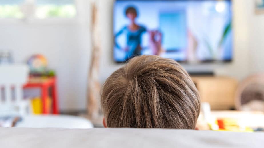 Die Kinderpraxis Ein Kind mit hellbraunem Haar sitzt in einem hellen, modernen Wohnzimmer auf einer Couch und blickt auf einen Fernseher, auf dem das verschwommene Bild einer Frau zu sehen ist. Kinderarzt Braunschweig Schlosscarree
