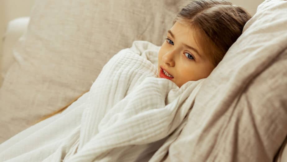 Die Kinderpraxis Ein junges Mädchen mit hellbraunem Haar liegt auf einer beigen Couch, eingewickelt in eine weiße Decke. Sie schaut mit neutralem Blick in die Kamera. Die Umgebung scheint ein Innenraum zu sein. Kinderarzt Braunschweig Schlosscarree