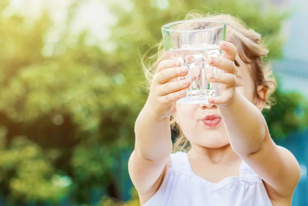 Die Kinderpraxis Ein kleines Kind in einem weißen ärmellosen Oberteil hält mit beiden Händen ein klares Wasserglas und streckt es nach vorne. Der Hintergrund ist ein Außenbereich mit grünem Laub und Sonnenlicht. Das Gesicht des Kindes ist leicht unscharf. Kinderarzt Braunschweig Schlosscarree