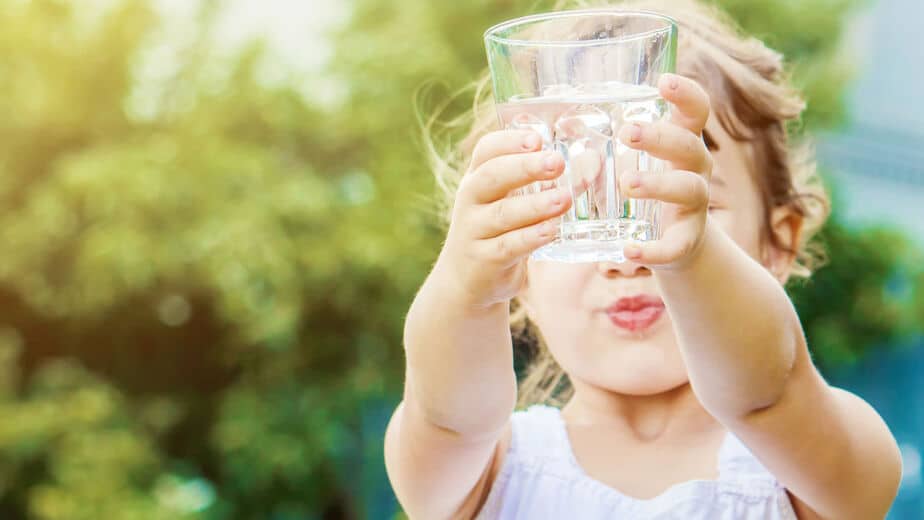Die Kinderpraxis Ein kleines Kind in einem weißen ärmellosen Oberteil hält mit beiden Händen ein klares Wasserglas und streckt es nach vorne. Der Hintergrund ist ein Außenbereich mit grünem Laub und Sonnenlicht. Das Gesicht des Kindes ist leicht unscharf. Kinderarzt Braunschweig Schlosscarree