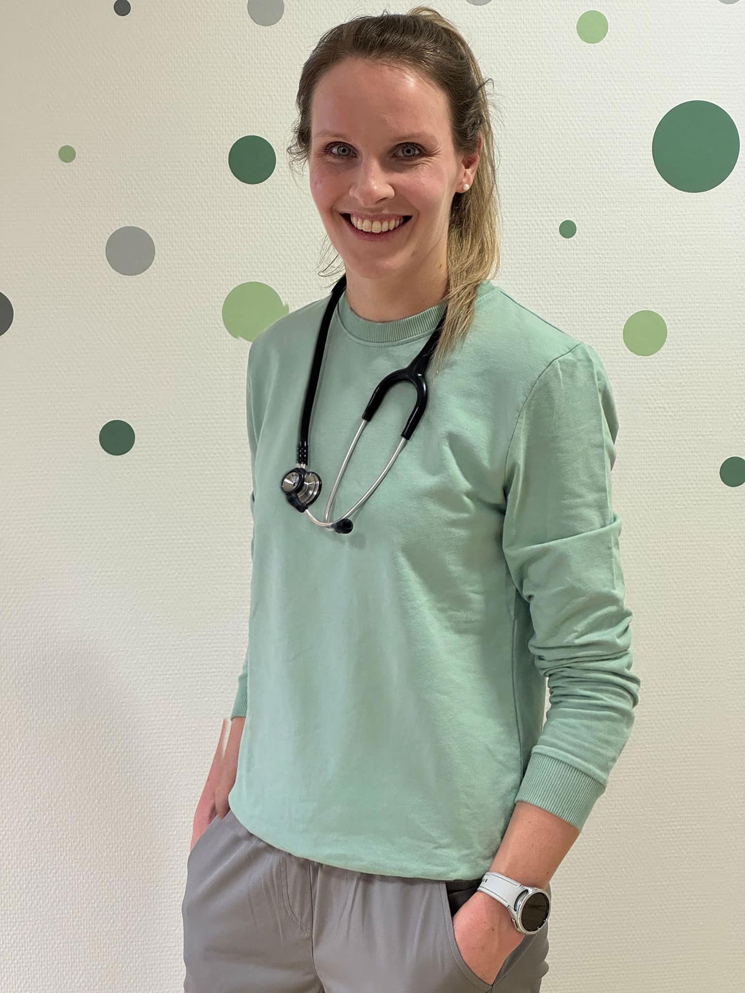 Die Kinderpraxis Eine Person, die ein mintgrünes langärmeliges Hemd, eine hellgraue Hose, eine Armbanduhr und ein Stethoskop um den Hals trägt, steht an einer mit grünen und grauen Punkten verzierten Wand und lächelt mit den Händen in den Taschen - ein einladendes Bild aus Über die Kinderpraxis. Kinderarzt Braunschweig Schlosscarree